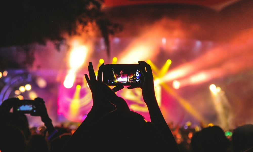 person holding smartphone taking video of a concert near stage with lights during nighttime
