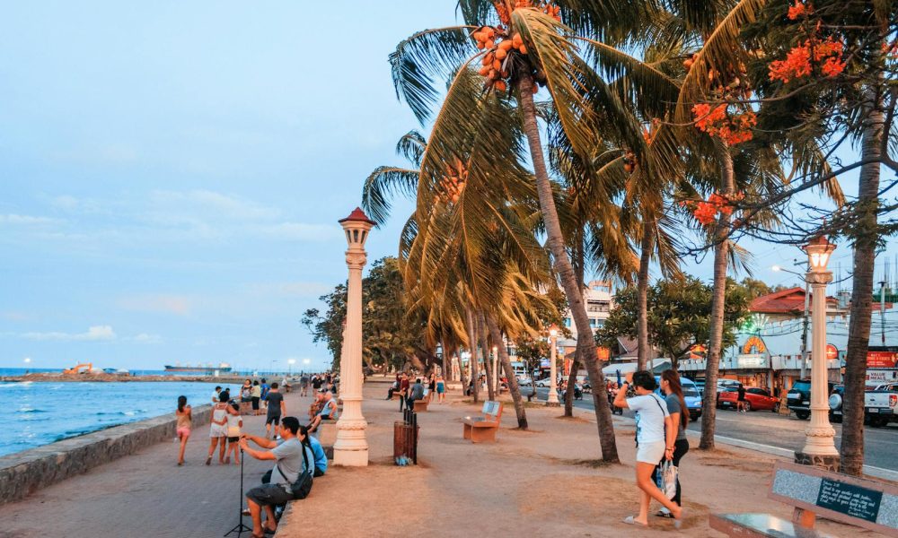 People stroll along a vibrant seaside promenade with palm trees and ocean views at dusk.