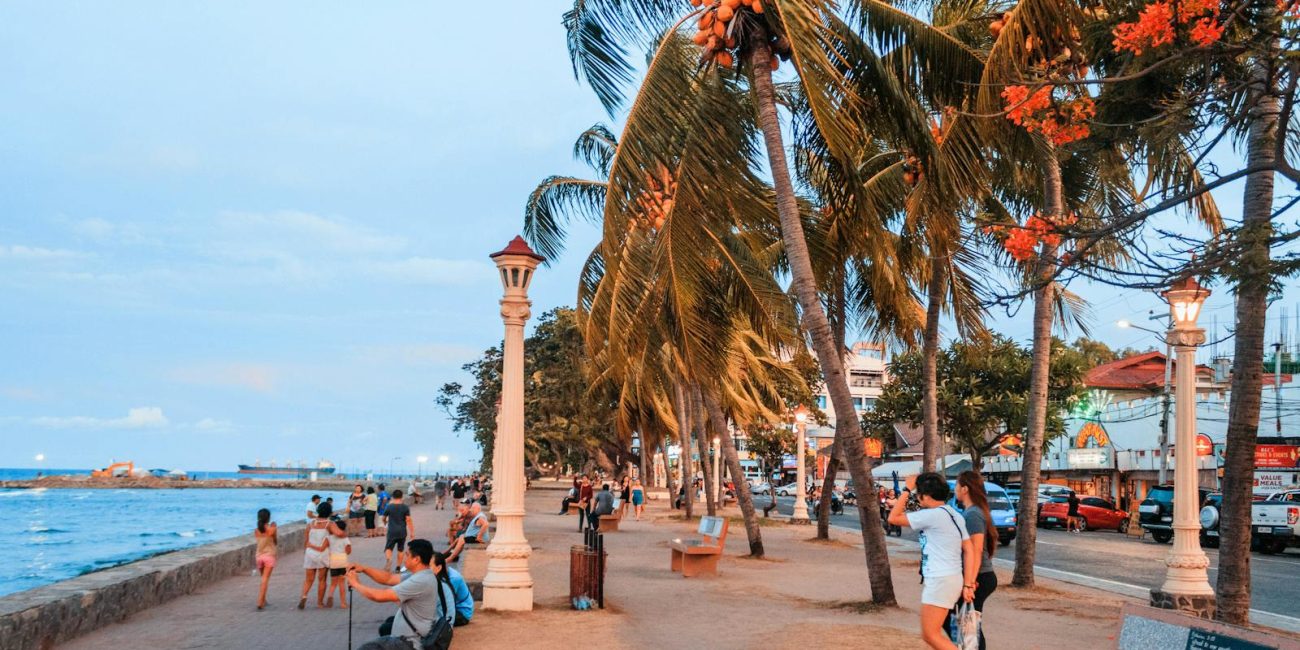 People stroll along a vibrant seaside promenade with palm trees and ocean views at dusk.