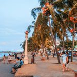 People stroll along a vibrant seaside promenade with palm trees and ocean views at dusk.