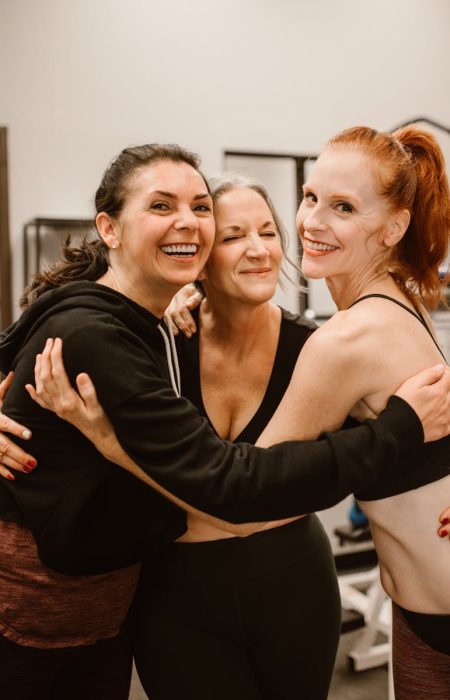 Three happy women hugging at the gym, celebrating friendship and wellness.