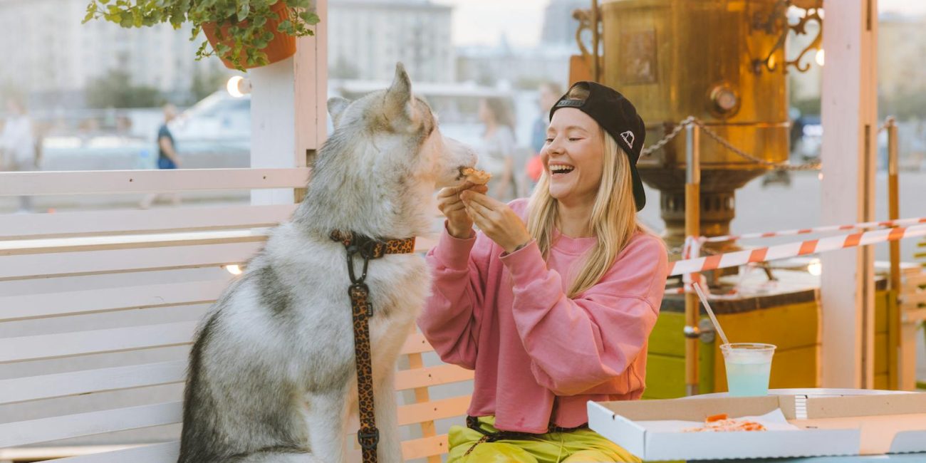 A joyful moment of a woman sharing pizza with her husky dog at an outdoor café.